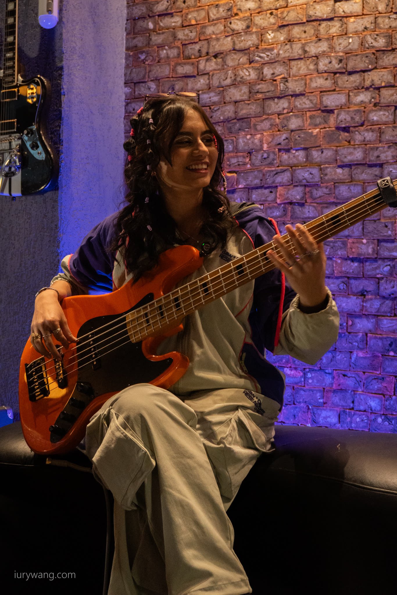 Baixista tocando e sorrindo, de corpo inteiro, diante da parede de tijolos