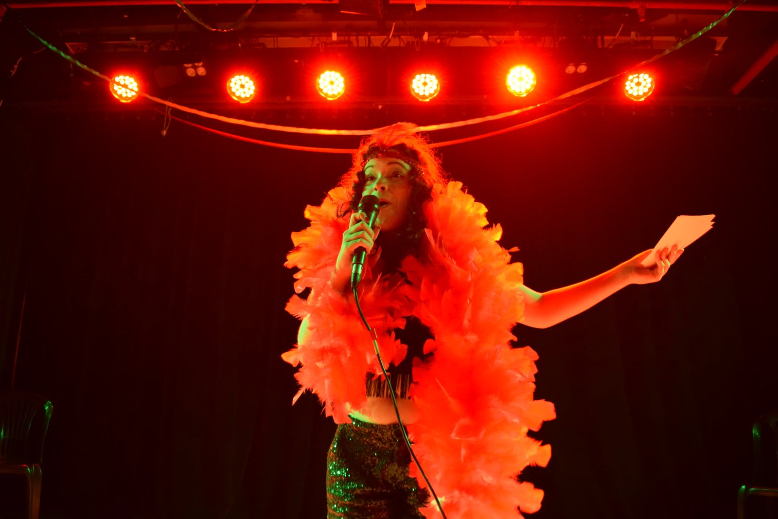 Atriz com boá de plumas laranja e microfone sob luz vermelha