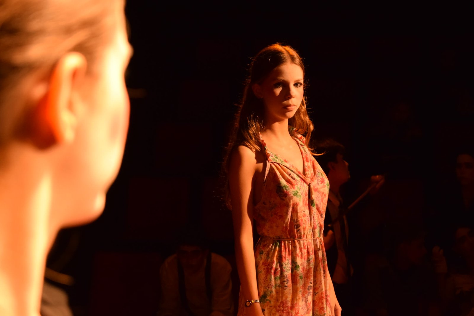 Atriz de vestido florido em cena, com outra atriz de perfil em primeiro plano