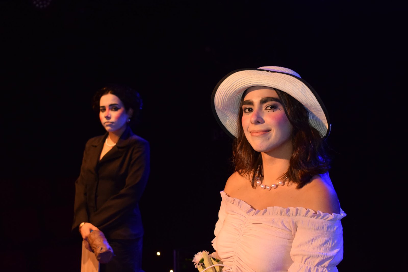 Atriz de blazer e atriz de chapéu branco com buquê em cena